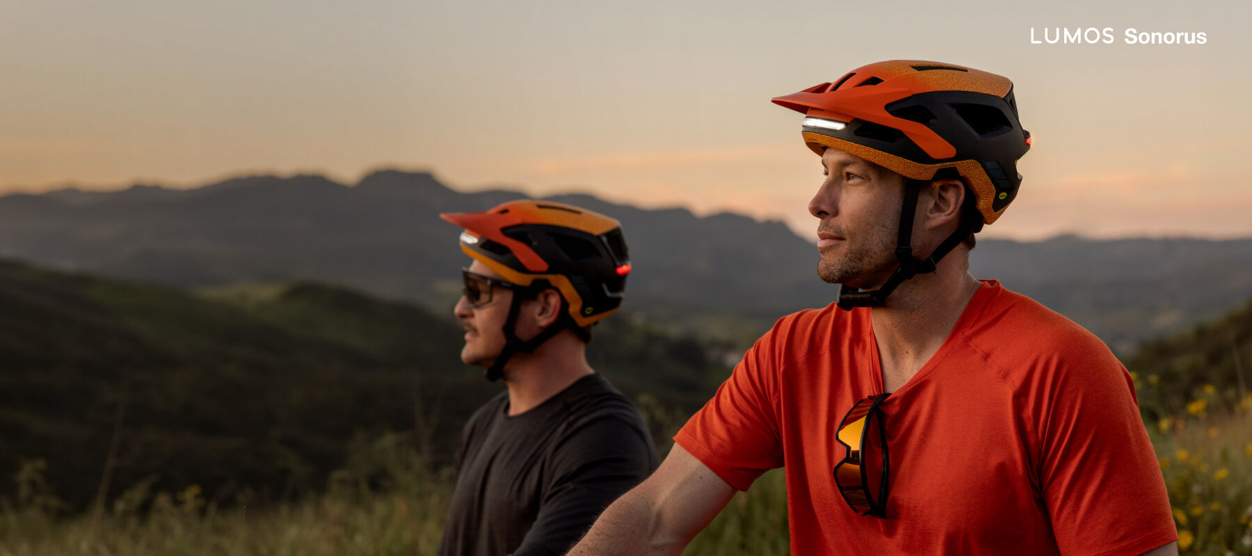 Two riders wearing orange Lumos Sonorus mesh intercom helmets in the hills at sunset